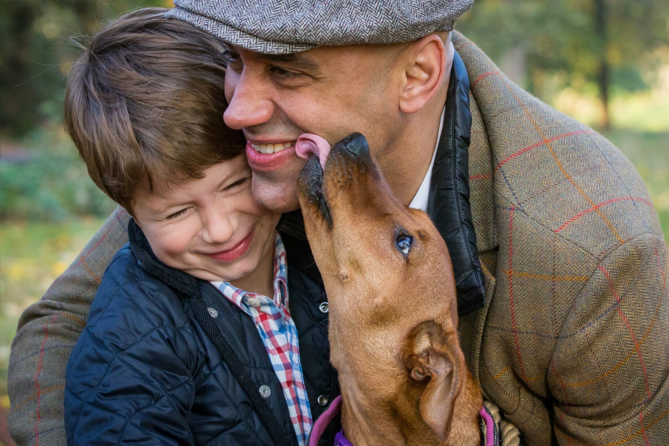 Outdoor family photograph of a boy and his dad laughing at the dog licking them