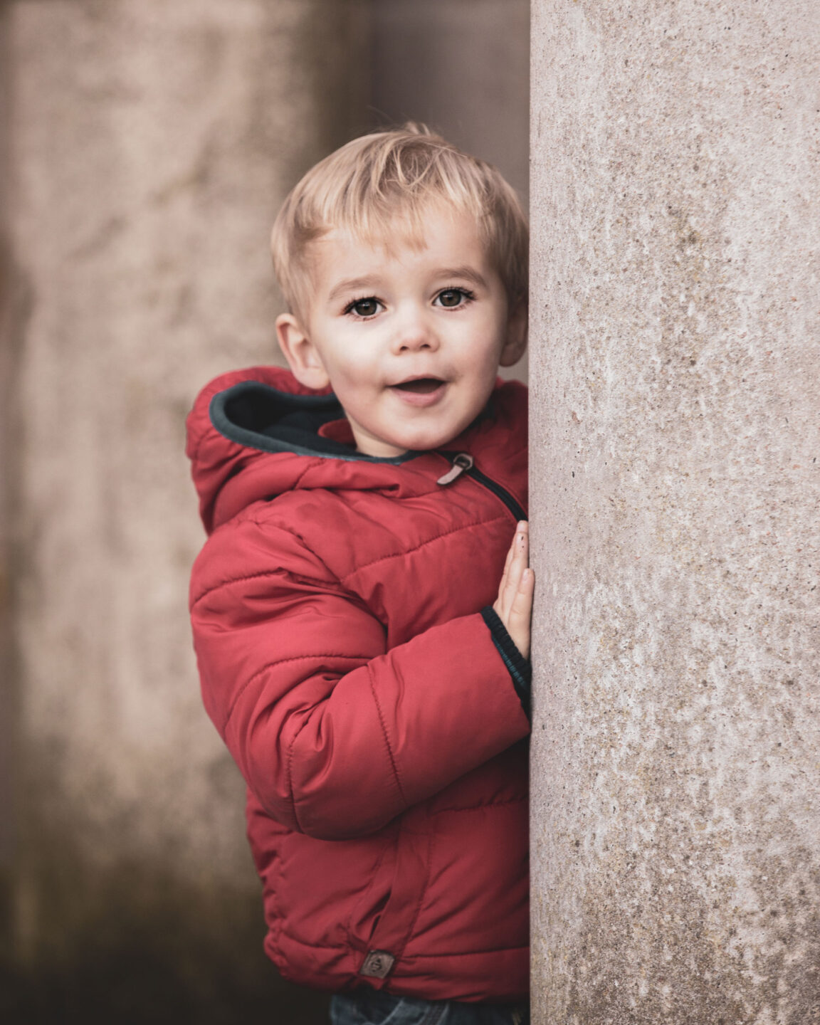 Cute blonde child peeking out from a pillar outdoors wearing a red coat
