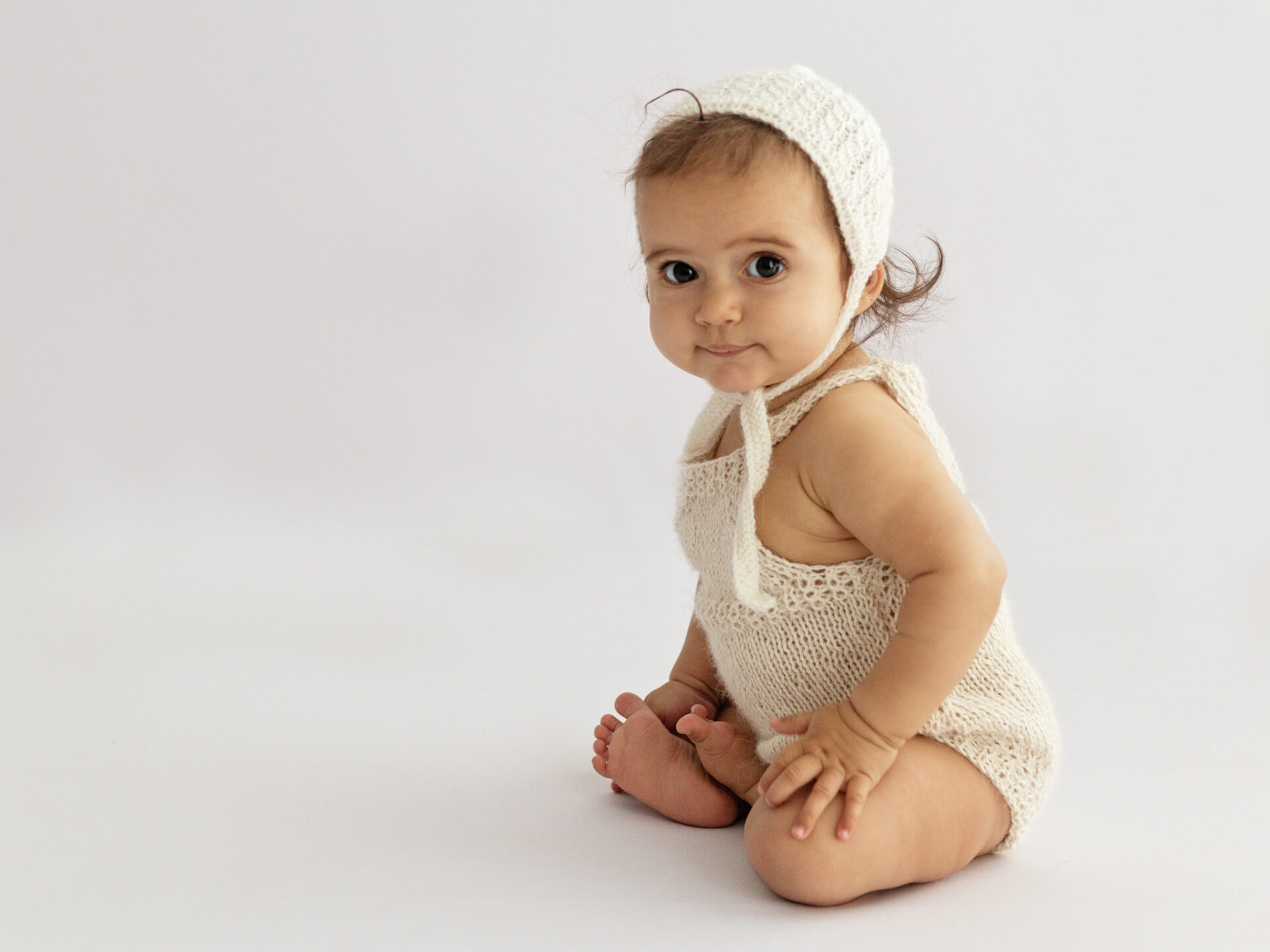Baby girl with big brown eyes sitting wearing knitted romper and bonnet